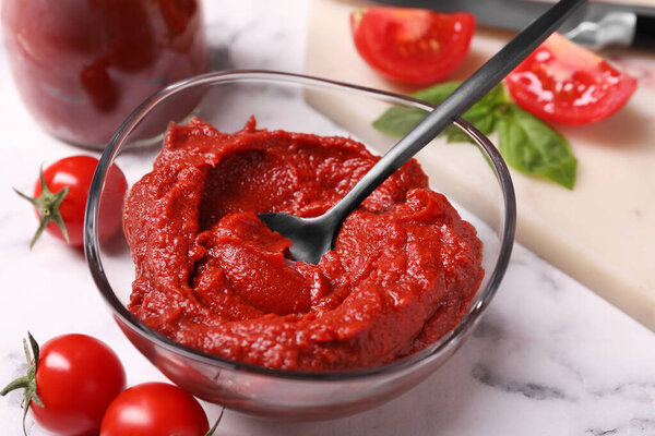 Стеклянная миска вкусной томатной пасты с ложкой и ингредиентами на белом мраморном столе, крупным планом