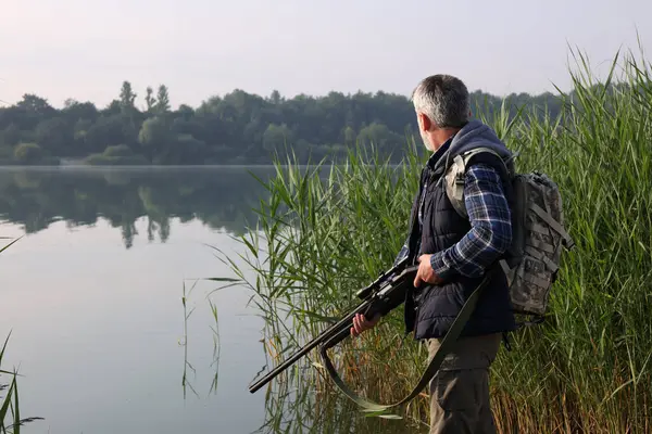 Man with hunting rifle near lake outdoors. Space for text