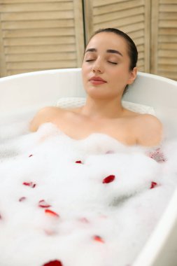 Kadın küvette köpük ve gül yapraklarıyla banyo yapıyor.