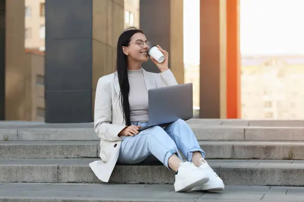 Happy young woman with cup of coffee using modern laptop on stairs outdoors