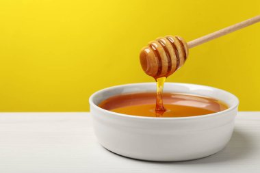 Pouring tasty honey from dipper into bowl at white wooden table against yellow background, closeup. Space for text clipart