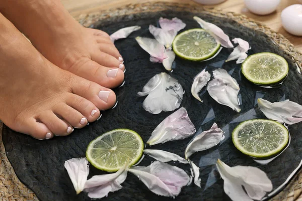 Woman soaking her feet in plate with water, flower petals and lime slices, closeup. Pedicure procedure
