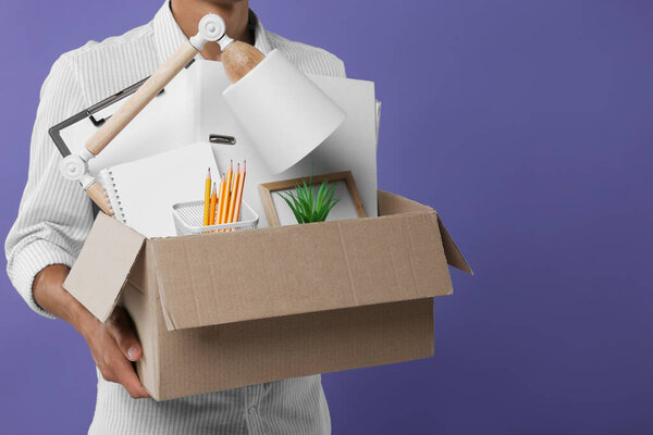 Unemployed young man with box of personal office belongings on purple background, closeup. Space for text