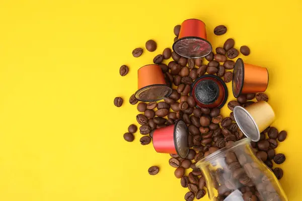Many coffee capsules, beans and glass jar on yellow background, flat lay. Space for text