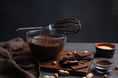 Bir kase çikolatalı krema, çırpı ve malzemeler gri masada.