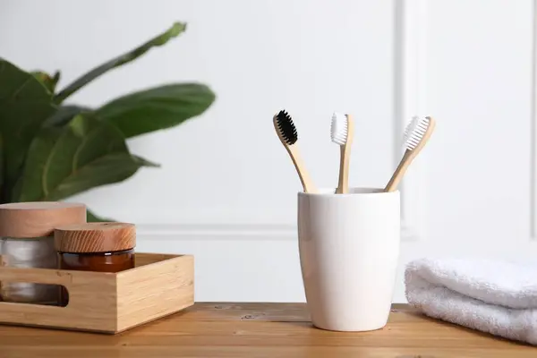 Bamboo toothbrushes in holder, towel, cosmetic products and leaves on wooden table