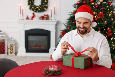 Noel Baba şapkalı genç adam dekore odasındaki masada Noel hediyesini açıyor.