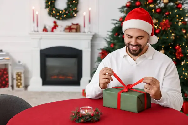 Счастливый Молодой Человек Шляпе Санты Открывающий Рождественский Подарок Столом Украшенной — стоковое фото