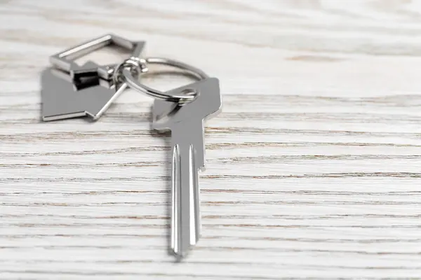 Key with keychain in shape of house on light wooden table, closeup. Space for text