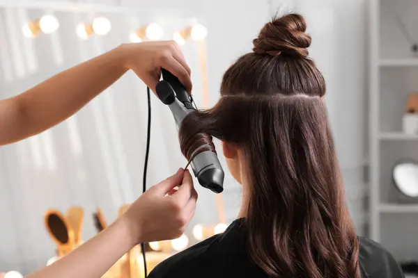 Hairdresser using curling hair iron while working with woman in salon, closeup
