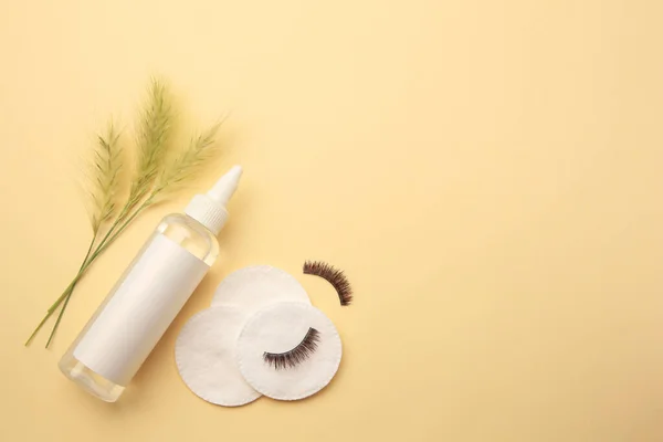 Bottle of makeup remover, cotton pads, false eyelashes and spikelets on yellow background, flat lay. Space for text