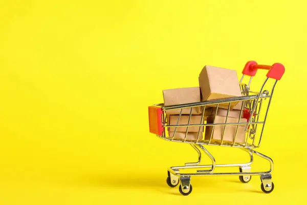 Small metal shopping cart with boxes on yellow background. Space for text