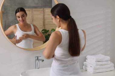 Güzel genç bir kadın banyoda aynanın yanında göğüslerini muayene ediyor.