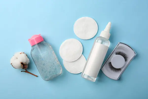 Bottles of makeup removers, cotton flower, pads and false eyelashes on light blue background, flat lay