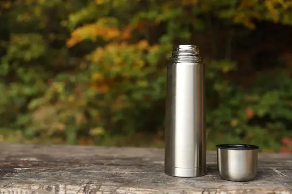 Metallic thermos and cup lid on wooden bench outdoors, space for text