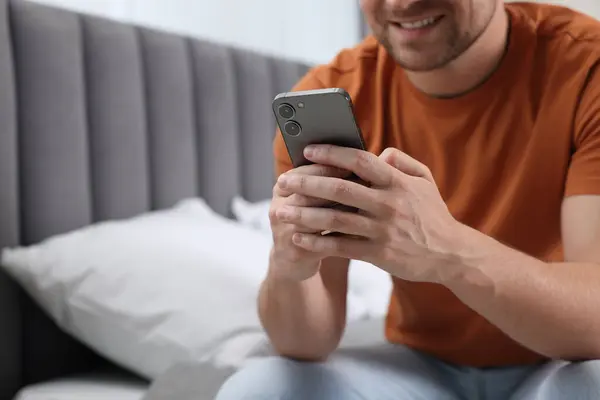 Man sending message via smartphone on bed at home, closeup. Space for text