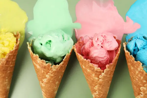 Melted ice cream in wafer cones on pale green background, flat lay