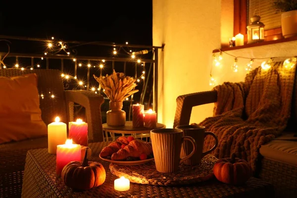 Rattan furniture, cups, fairy lights, burning candles and other autumn decor on outdoor terrace at night