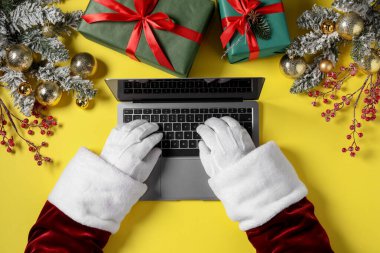 Noel Baba dizüstü bilgisayar kullanıyor, yakın plan. Hediye kutuları ve sarı arka planda Noel dekoru, üst manzara