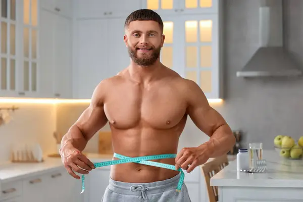 Athletic young man measuring his waist with tape in kitchen. Weight loss