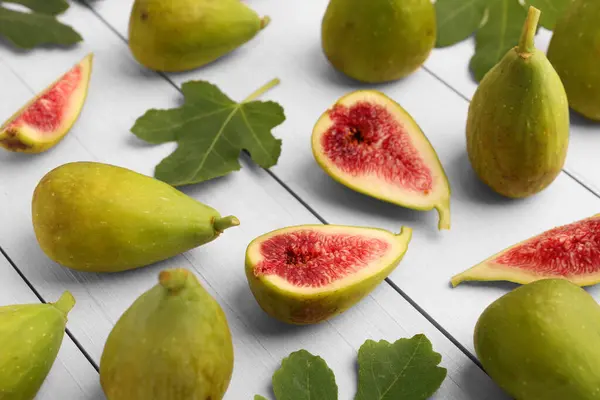 Cut and whole green figs with leaves on white wooden table, closeup