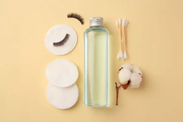 Bottle of makeup remover, cotton flower, pads, swabs and false eyelashes on yellow background, flat lay