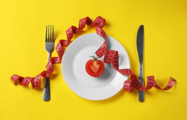Yarım tabak domates, çatal bıçak takımı ve sarı arka planda mezura. Diyet kavramı
