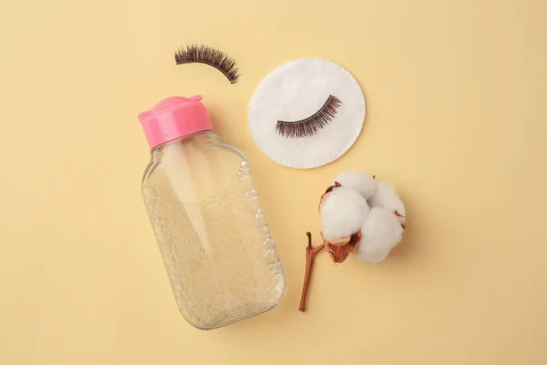 Bottle of makeup remover, cotton flower, pad and false eyelashes on yellow background, flat lay