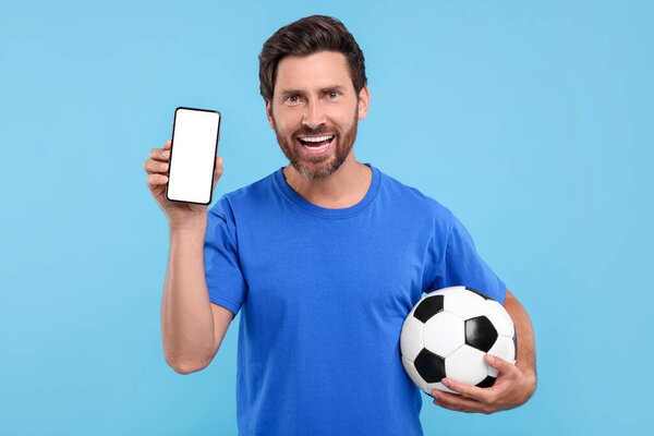 Happy sports fan with soccer ball and smartphone on light blue background