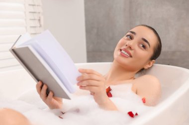 Küvette köpük ve gül yapraklarıyla banyo yaparken kitap okuyan mutlu kadın.