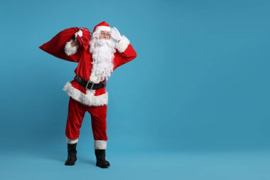 Noel Baba bir çanta dolusu hediyeyle açık mavi arka planda poz veriyor.