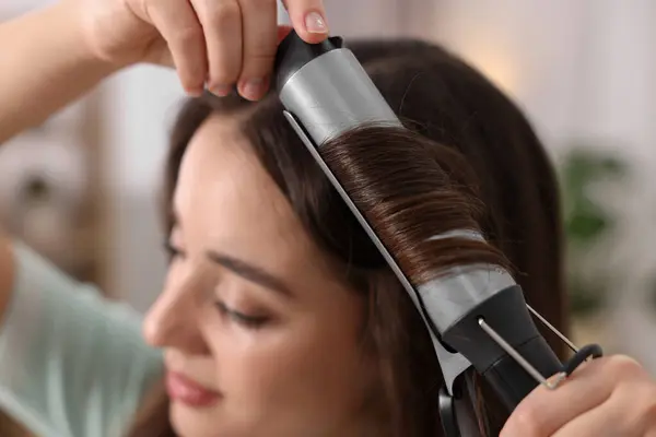 Woman using curling hair iron indoors, selective focus