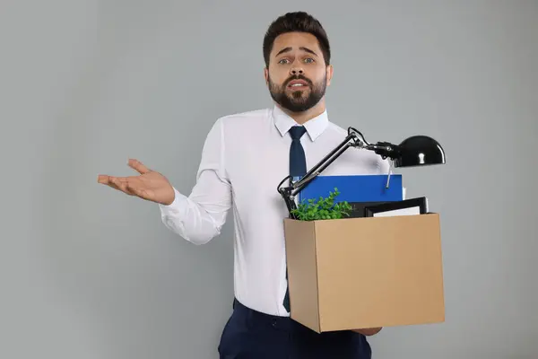 Confused unemployed man with box of personal office belongings on light grey background