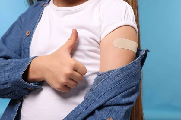 Girl with sticking plaster on arm after vaccination showing thumbs up against light blue background, closeup
