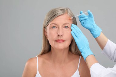 Doktor, açık gri arka planda yaşlı kadına yüz enjeksiyonu yapıyor. Yakın plan. Estetik ameliyat