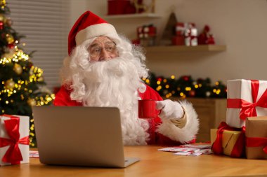 Noel Baba elinde içki bardağıyla masada dizüstü bilgisayar ve odada Noel hediyeleri tutuyor.