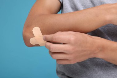Man putting sticking plasters onto elbow on light blue background, closeup clipart