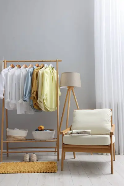 Wardrobe organization. Rack with different stylish clothes, armchair and lamp near grey wall indoors