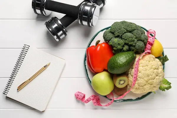 Healthy diet. Plate with products, measuring tape, dumbbells and notebook on white wooden table, flat lay