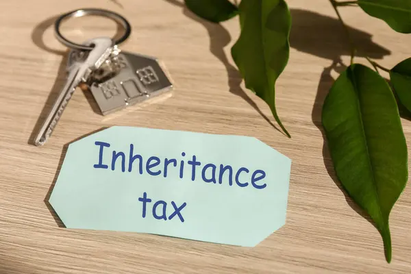Inheritance Tax. Card, key with key chain in shape of house and green leaves on wooden table, closeup
