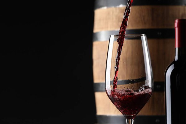 Pouring red wine into glass near wooden barrel against black background. Space for text