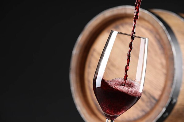 Pouring red wine into glass near wooden barrel against black background, closeup. Space for text