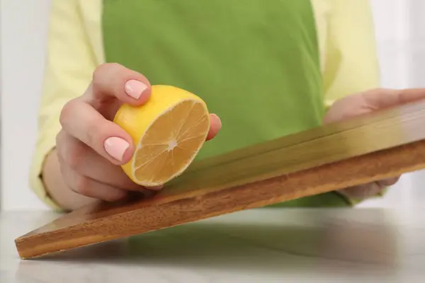 Woman rubbing wooden cutting board with lemon at white table, closeup