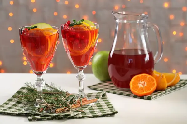 Christmas Sangria cocktail in glasses and jug, ingredients and fir tree branch on white table against blurred lights