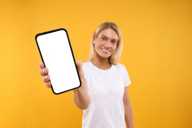 Turuncu arka planda boş ekranı olan akıllı telefonu tutan mutlu kadın, seçici odaklanma