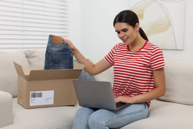 Evdeki kanepede dizüstü bilgisayar kullanarak yeni açılmış kotları olan genç bir kadın. İnternetten alışveriş