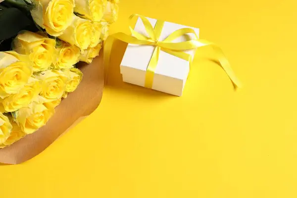 Beautiful bouquet of roses and gift box on yellow background, above view. Space for text
