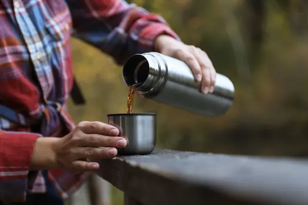 Woman pouring hot drink from metallic thermos into cup lid outdoors, closeup