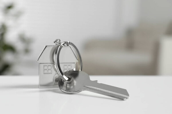 Key with keychain in shape of house on white table against blurred background, closeup. Space for text