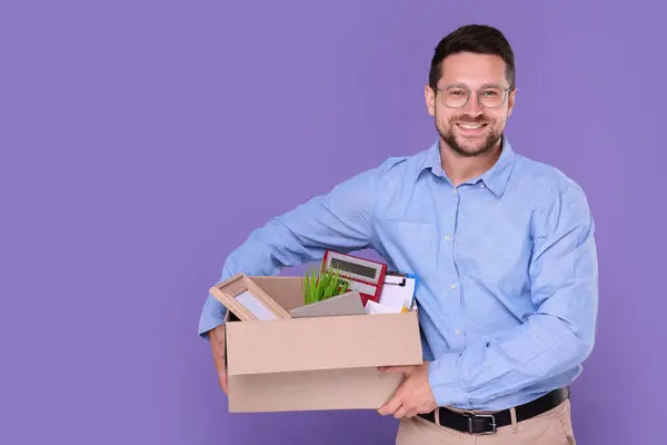 Happy unemployed man with box of personal office belongings on purple background. Space for text